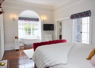 Bright bedroom in Bunbury featuring a white bed with a red velvet headboard, wall-mounted TV, and a freestanding bathtub by a large arched window—perfect for a luxurious retreat flooded with natural light.