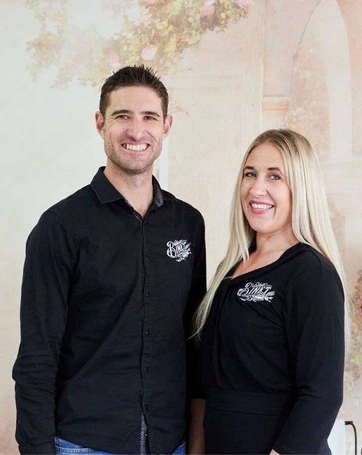 A man and a woman stand side by side, both smiling at the camera, wearing black shirts with matching embroidered logos, against a soft, floral background. A man and a woman stand side by side, both smiling at the camera, wearing black shirts with matching embroidered logos, against a soft, floral background.