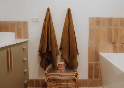 Two brown towels hang on hooks above a wooden stool in a bathroom with beige tiles, olive green drawers, and a white bathtub—creating the perfect ambiance for a luxurious Parent Retreat Bunbury.