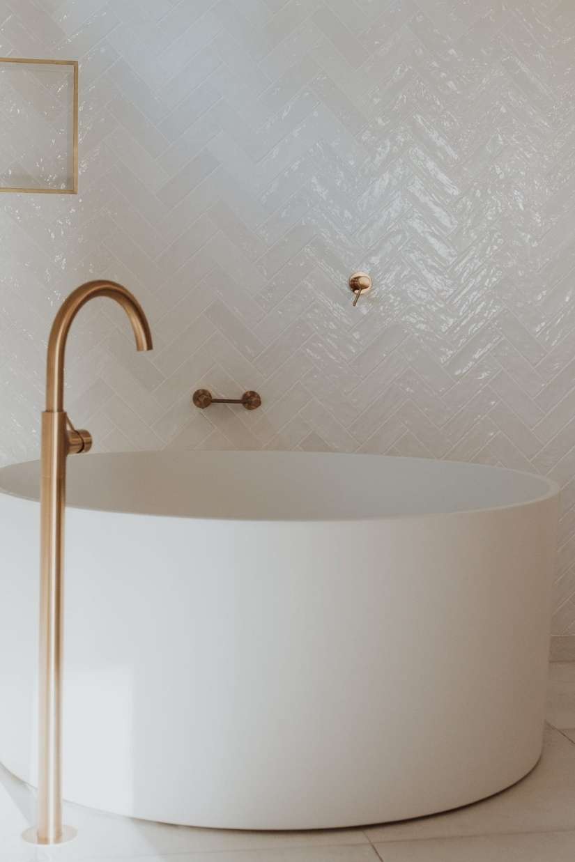 A modern white freestanding bathtub with a tall gold faucet is set against a glossy white herringbone tiled wall. A modern white freestanding bathtub with a tall gold faucet is set against a glossy white herringbone tiled wall.