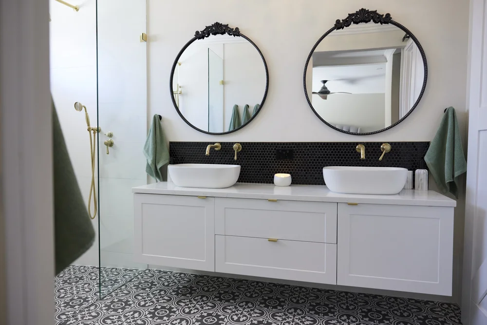 Modern bathroom with two white sinks, round mirrors, gold fixtures, green towels, black hexagonal tile backsplash, and patterned tile floor—perfect inspiration for your next Bathroom Renovations in Bunbury. Modern bathroom with two white sinks, round mirrors, gold fixtures, green towels, black hexagonal tile backsplash, and patterned tile floor—perfect inspiration for your next Bathroom Renovations in Bunbury.