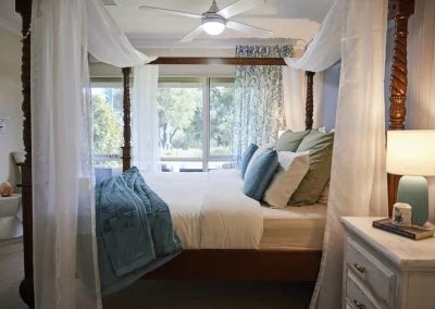 A luxurious parent retreat in Bunbury, this bedroom features a four-poster bed with white and blue bedding, sheer canopy curtains, a nightstand with lamp and book, and large windows letting in natural light.