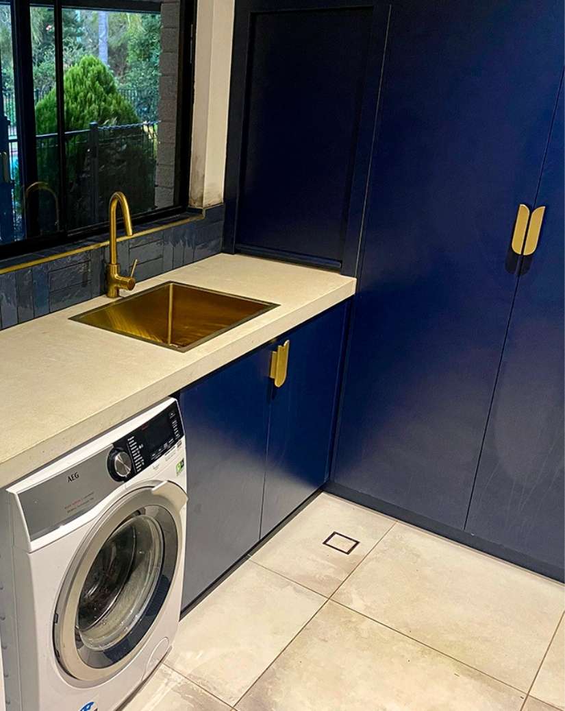 A modern laundry room with blue cabinets, a white front-loading washing machine, a beige countertop, and a gold sink faucet beneath a window. A modern laundry room with blue cabinets, a white front-loading washing machine, a beige countertop, and a gold sink faucet beneath a window.
