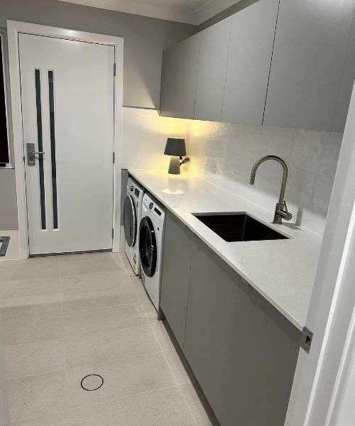 Modern laundry room with grey cabinets, white countertop, a sink, washing machine, dryer, and a small lamp near the backsplash under-cabinet lighting. Modern laundry room with grey cabinets, white countertop, a sink, washing machine, dryer, and a small lamp near the backsplash under-cabinet lighting.