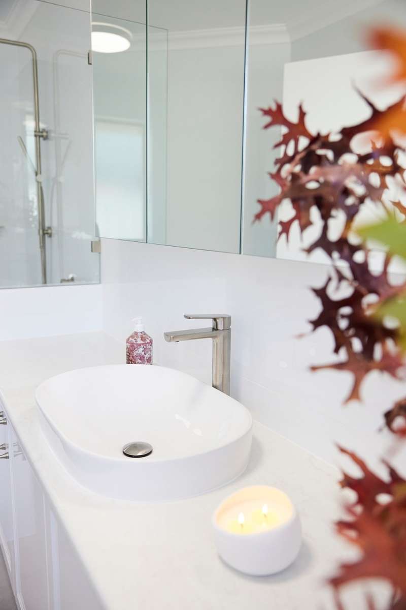 Modern bathroom countertop with a white vessel sink, silver faucet, candle, soap dispenser, and autumnal leaves in the foreground. A shower area is visible in the background. Modern bathroom countertop with a white vessel sink, silver faucet, candle, soap dispenser, and autumnal leaves in the foreground. A shower area is visible in the background.