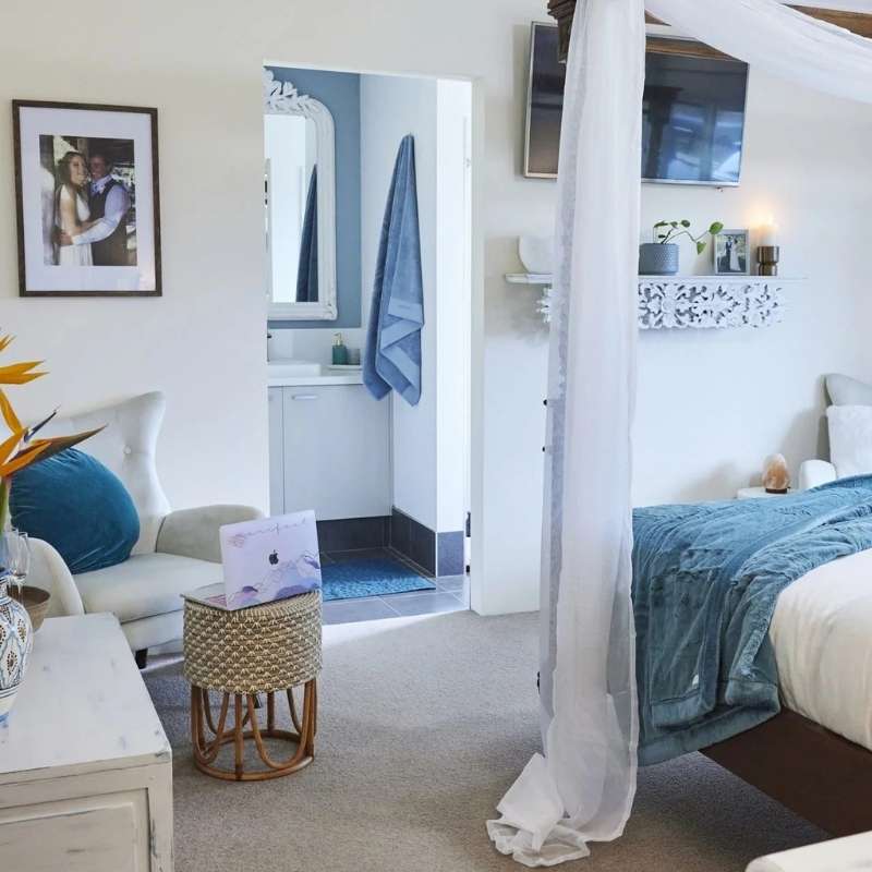 A cozy bedroom with a canopy bed, blue bedding, a small table, two chairs, a framed photo on the wall, and an open doorway leading to a bathroom with a blue towel. A cozy bedroom with a canopy bed, blue bedding, a small table, two chairs, a framed photo on the wall, and an open doorway leading to a bathroom with a blue towel.
