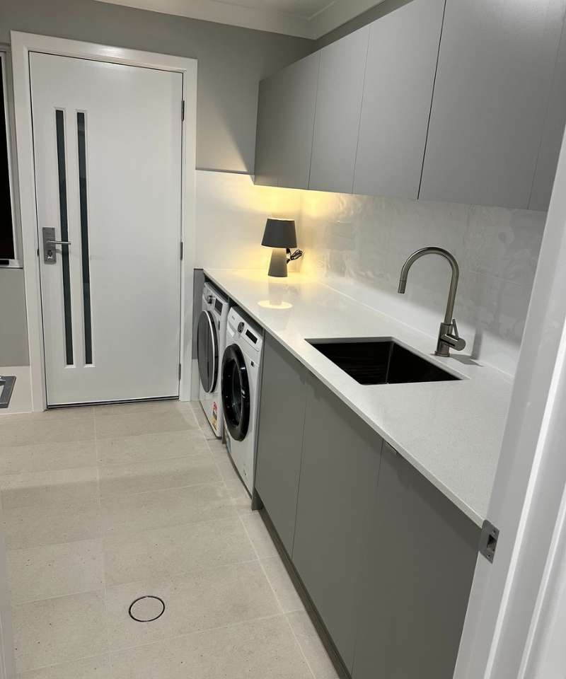 Modern laundry room with grey cabinets, a white countertop, built-in sink, front-loading washer and dryer, and a small table lamp near the back wall. Modern laundry room with grey cabinets, a white countertop, built-in sink, front-loading washer and dryer, and a small table lamp near the back wall.