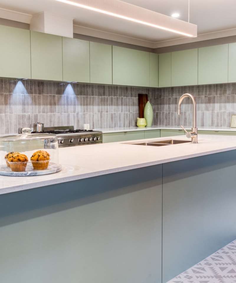Modern kitchen with light green cabinets, a built-in stovetop, tiled backsplash, and a countertop featuring two muffins on a glass stand next to a sink with a gooseneck faucet. Modern kitchen with light green cabinets, a built-in stovetop, tiled backsplash, and a countertop featuring two muffins on a glass stand next to a sink with a gooseneck faucet.