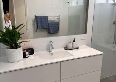Modern Dalyellup bathroom featuring a white vanity, sink, potted plant, candle, soap dispenser, towel rack with blue towels, and a sleek glass shower enclosure—perfect inspiration for your next bathroom renovation.